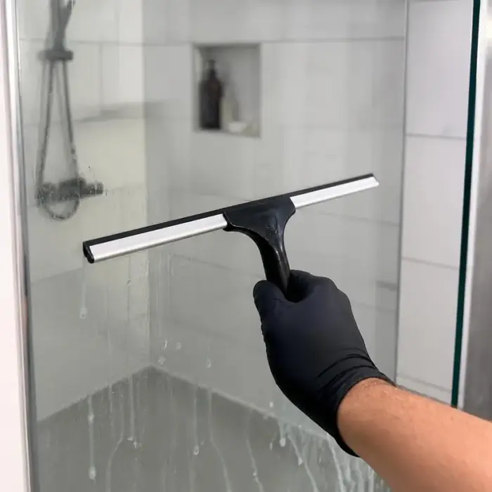 Gloved hand pulling a rubber squeegee down a wet glass shower door during a one-time deep cleaning